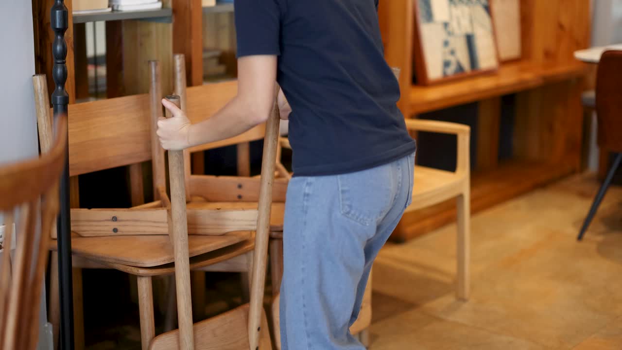 Individual arranges wooden chairs in a casual cafe, natural lighting, steady handheld camera movement