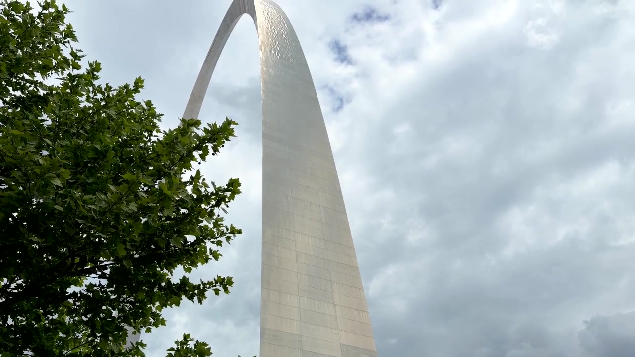 Una panoramica verso l'alto rivela l'imponente Gateway Arch a St. Louis, Missouri, incorniciato da alberi verdi e nuvole spettacolari