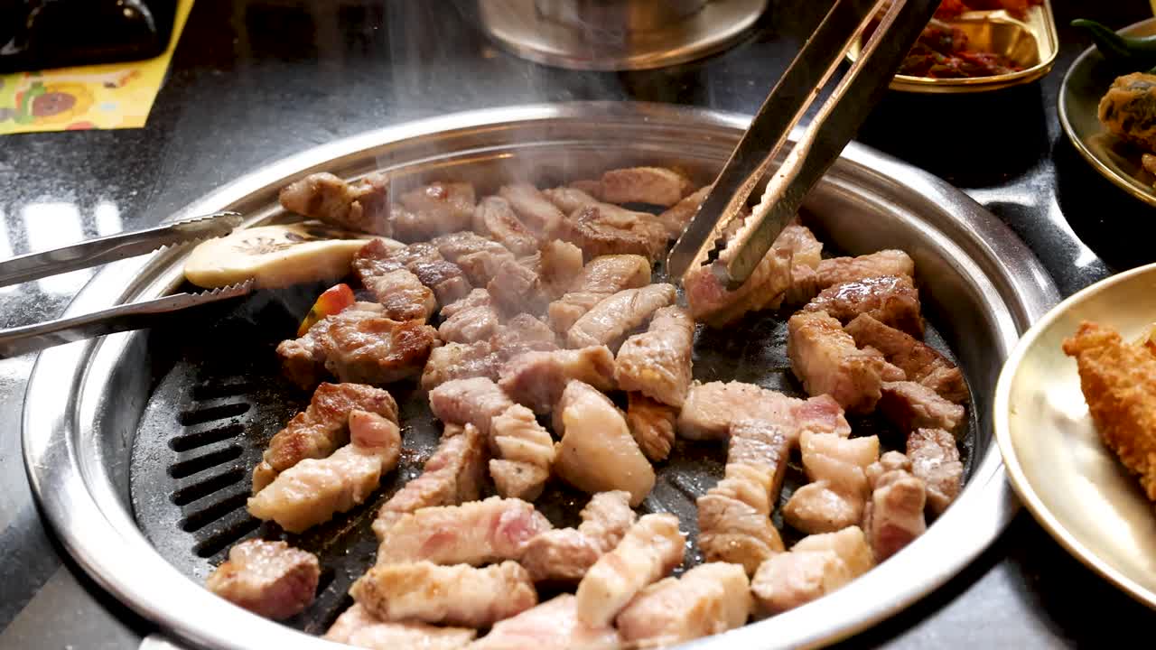 Juicy pork belly grilling on tabletop Korean BBQ, steam rising as tongs turn the meat
