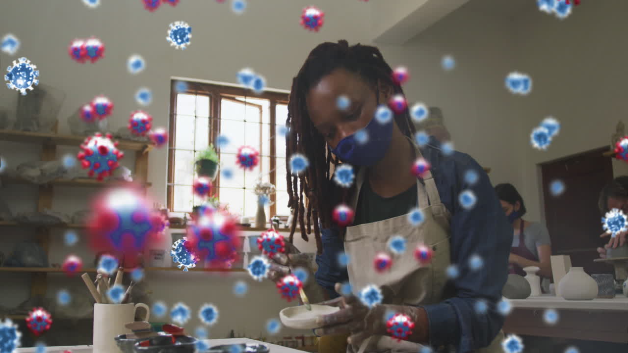 Animation of covid 19 cells over women and man working in pottery studio wearing face masks