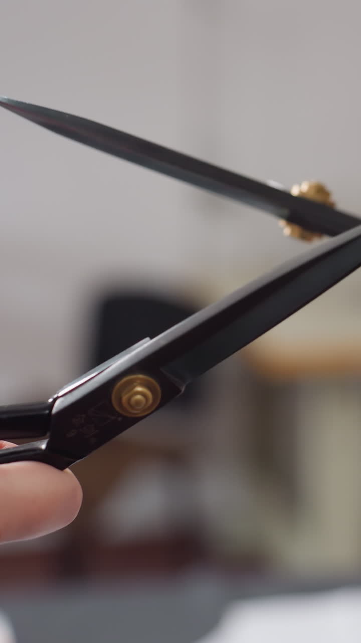 Primer plano de una mano sujetando unas tijeras negras, cortes de precisión en un taller de estudio, detalle dorado en el pivote, inspección de la cuchilla, artesano probando el filo y ajustando el tornillo, movimiento concentrado de la mano, corte lento y deliberado