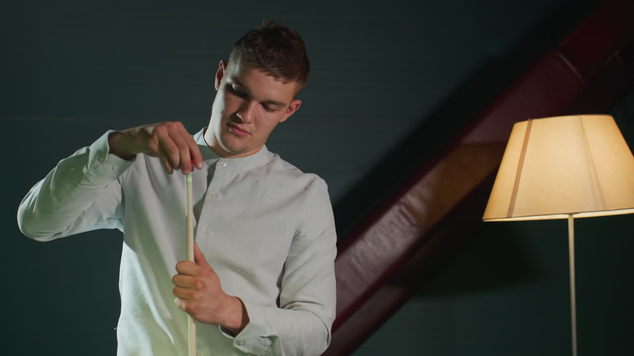 어두운 배경에  셔츠를 입은 젊은이의 전면 뷰, 큐 스틱을 조심스럽게 들고, 집중된 표정으로 빨간 개를 적용합니다. 따뜻한 조명은 집중력을 강조합니다.