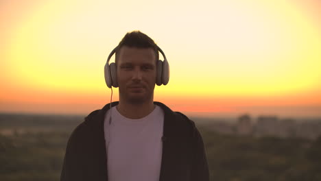 smiling and laughing handsome young man listening music from his smartphone in wireless headphones. in slow motion a man stands on the roof at sunset and looks at the beautiful view from the height of the city