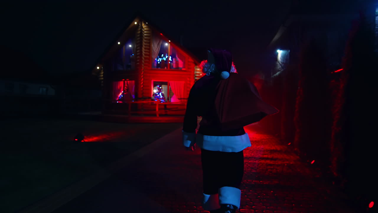 Santa Claus coming up to the cozy wooden house with bright Christmas lights. Beautiful wooden house highlighted with red light. Santa's visit on the late evening time.