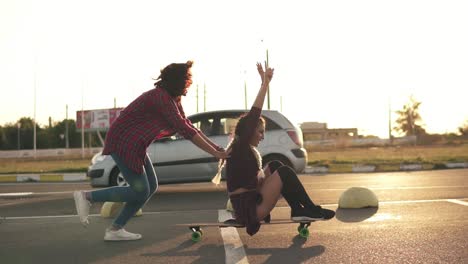 vista lateral de una mujer con las manos levantadas sentada en un longboard mientras su amiga la empuja hacia atrás y corre durante la puesta de sol