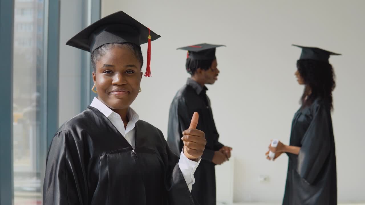One young African-American woman stands in the foreground and looks at the camera with a diploma in her hands. Young African American man and woman communicate with each other in the background.