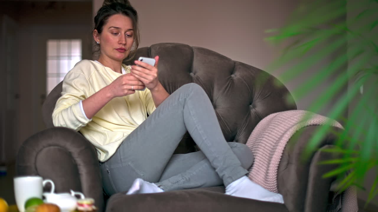 Caucasian woman sitting on a chair is on her smartphone. Table with tea and fruits nearby