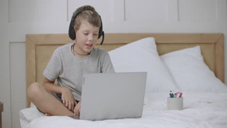 little boy is drawing lying on bed in his room and communicating online by web camera and headphones