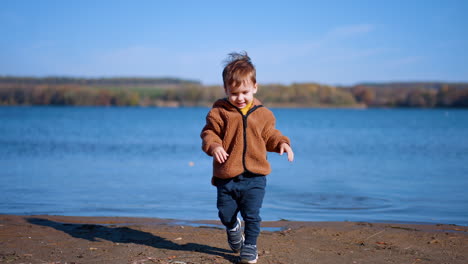 lovely cute kid in warm brown jacket approaches river. happy child throws the stone and goes for the new stone.