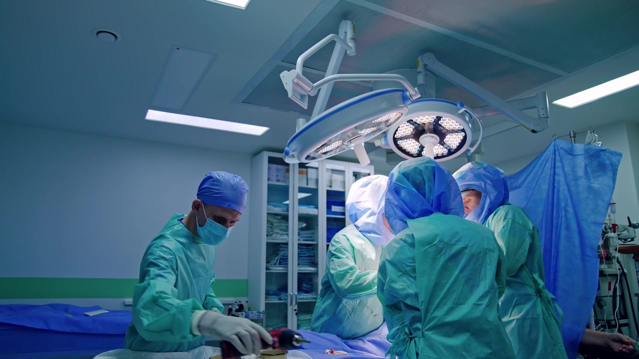 Male assistant arranges the tools on the surgical stand. Team of doctors in protective helmets work at operational table.