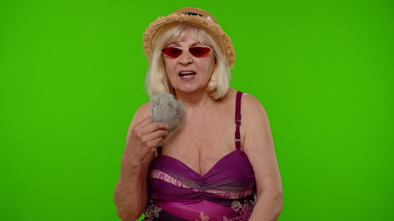 성숙한 할머니 여행자는 복권에서 승리하여 기하고, 돈, 달러, 현금을 흔들며, 미소 짓고 있습니다.