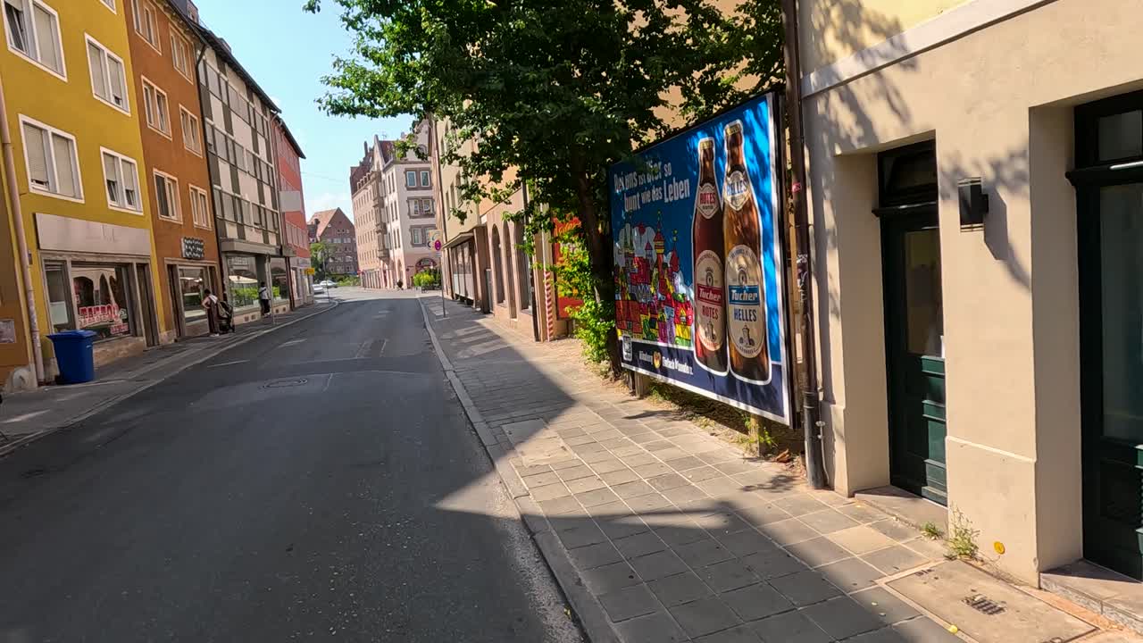 A steady camera moves along a quiet, sunlit street in Nuremberg, Germany, passing colorful storefronts, advertisements, and trees on a clear summer day