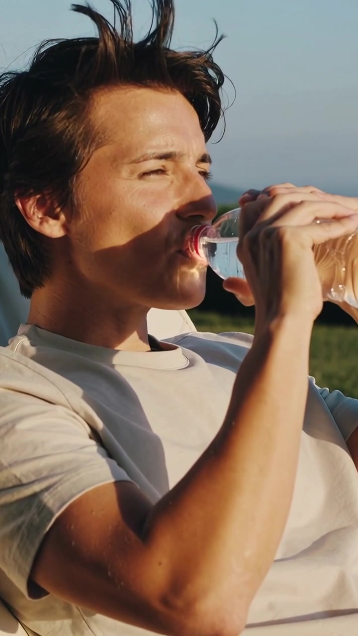 Vertical video: Sipping man on lawn recliner in light tee raising clear bottle pink cap sun urging