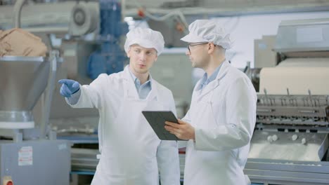 two young food factory employees discuss work-related matters. male technician or quality manager uses a tablet computer for work. they wear white sanitary hat and work robes.