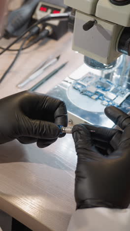 una vista cercana de un técnico trabajando en una placa de circuito bajo un microscopio, el técnico, con guantes negros, está girando un tornillo en su mano varias herramientas son visibles en el escritorio