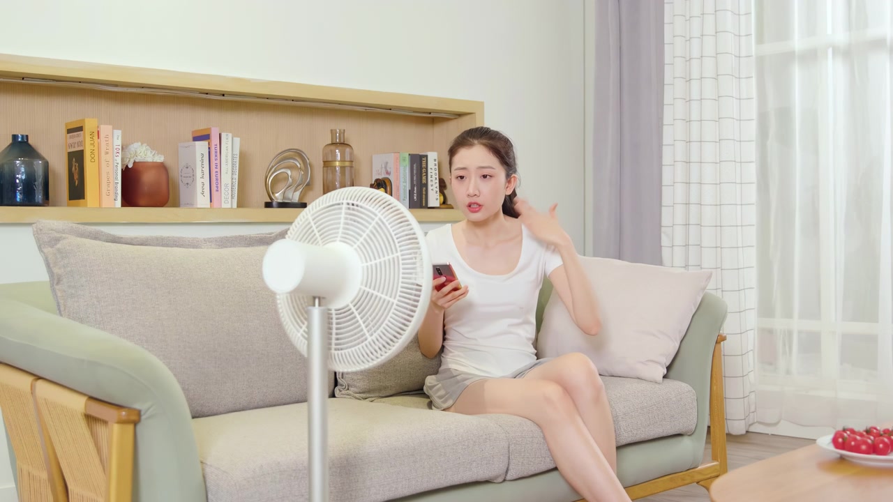 Portrait of young girl sitting on cozy couch sofa, uses a smartphone while cooling down in front of an electric fan, escapes from the heat during summer hot weather apartment