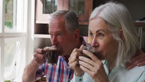 Älteres-Paar-Trinkt-Gemeinsam-Kaffee,-Während-Es-Zu-Hause-In-Der-Küche-Aus-Dem-Fenster-Schaut