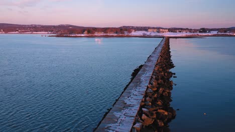 Luftaufnahmen,-Die-Bei-Sonnenaufgang-Diagonal-über-Einen-Schneebedeckten-Felsigen-Wellenbrecher-In-Rockland-Maine-Fliegen