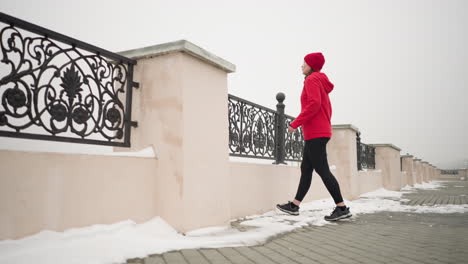 mujer con capucha roja y leggings negras se mueve a la valla de hierro y descansa las manos en la barandilla decorativa de metal, el suelo nevado que la rodea, el camino urbano que conduce a la distancia, la atmósfera de invierno