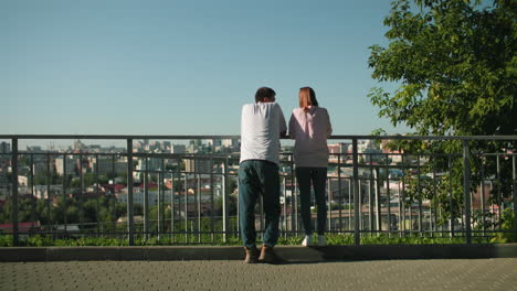 siblings leaning on iron railing having a warm interaction with the girl smiling at her brother, background features a vibrant cityscape with trees and sunlight