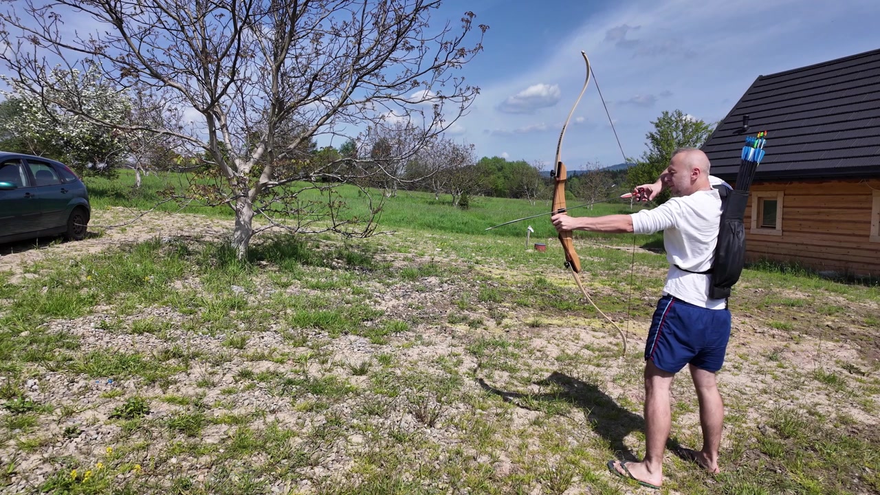 Outdoor archery session featuring a man drawing, releasing and preparing the next arrow. Beautiful countryside, wooden cabin, clear blue sky and active lifestyle.