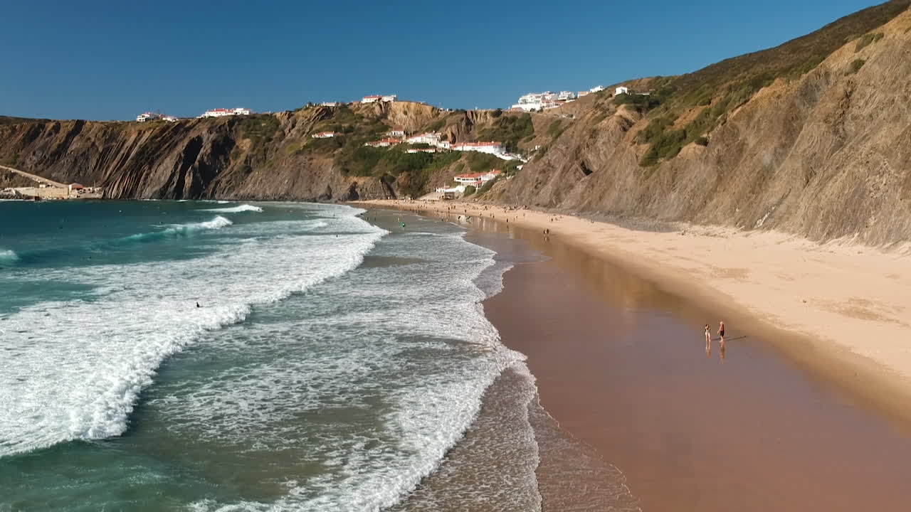 Imagem aérea de drone de ondas de espuma a quebrar numa praia junto a algumas casas num penhasco no Algarve, Portugal.