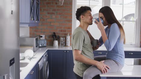 feliz pareja biracial sentada en el mostrador en la cocina, bebiendo café