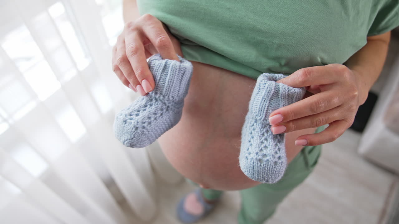 Future mommy stands at the window holding little baby booties on her bare stomach. She makes tiny steps by the booties on her belly. Mother puts one hand carefully on her tummy close up.