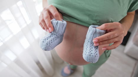 future mommy stands at the window holding little baby booties on her bare stomach. she makes tiny steps by the booties on her belly. mother puts one hand carefully on her tummy close up.