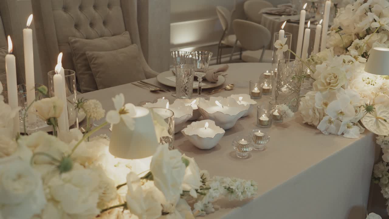 Allestimento di lusso per tavolo nuziale con candele, bicchieri e decorazioni floreali bianche in un interno di ricevimento romantico.