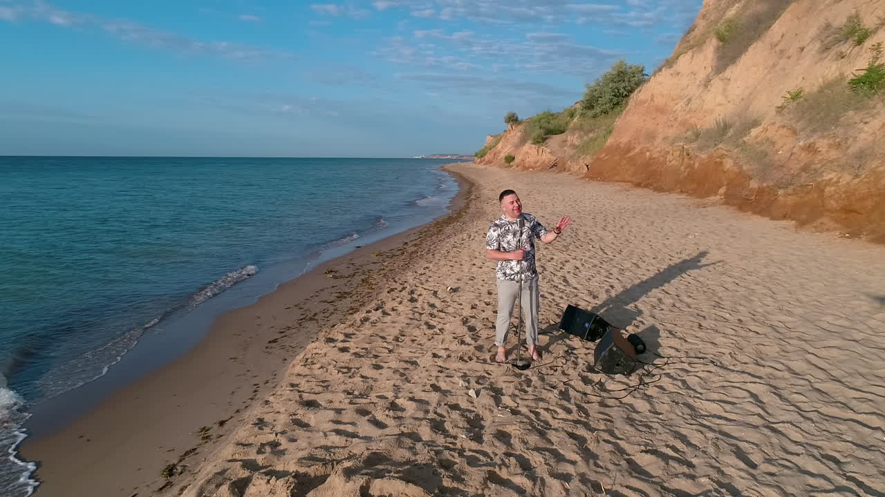Man singer on a sea beach. Musician man singing into microphone while standing on a sand shore on blue sea background. Orbital view.