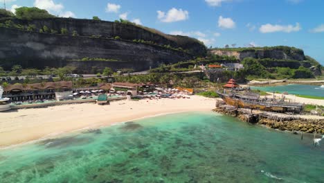 Minoo-Beach-Club-Orilla-De-Arena-Blanca-Con-Mar-Turquesa-Tranquilo-En-Uluwatu-Bali,-Playa-Melasti-Con-Acantilados-Escarpados-En-El-Fondo---Toma-Aérea-De-Retroceso