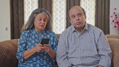 Old-Indian-husband-watching-TV,-wife-busy-with-her-phone