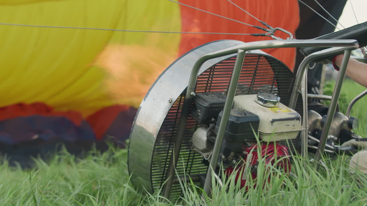 Person igniting hot air balloon burner as engine fan propels powerful flame into colorful nylon envelope on grassy field during windy outdoor launch setup with tension cables stretching across frame