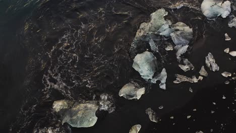 Waves-of-Ocean-crashing-against-icy-icebergs-lying-on-black-sandy-beach-in-Iceland