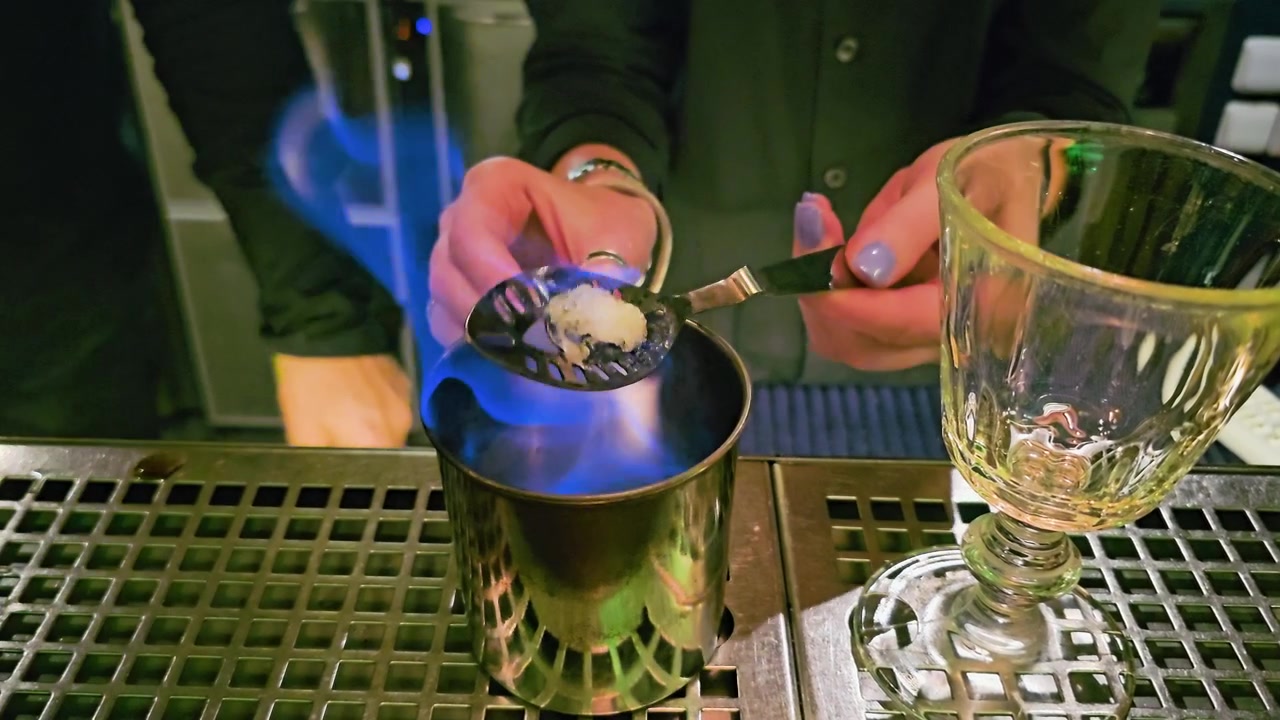 Scena suggestiva di un cocktail bar che mostra il servizio di assenzio con fiamma blu, zolletta di zucchero, cucchiaino forato, tazza di metallo, calice di vetro, mani del barista e vassoio raccogligocce riflettente in acciaio inossidabile di notte