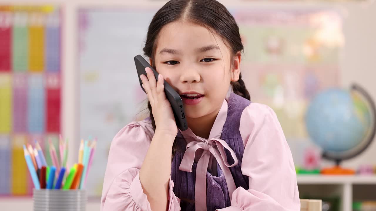 Smiling young girl talks on smartphone at desk in brightly lit, colorful classroom environment