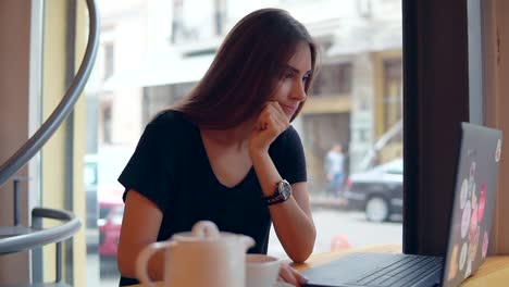 Young-woman-sitting-in-the-coffee-shot-by-the-window-having-a-coffee-break,-she-is-using-her-laptop,-working-and-surfing-in-the