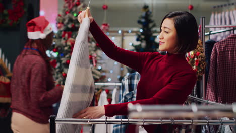 asian client browsing through rack of clothes in christmas themed shop, checking for fitting blouse size. woman in xmas ornate fashion boutique during festive holiday season