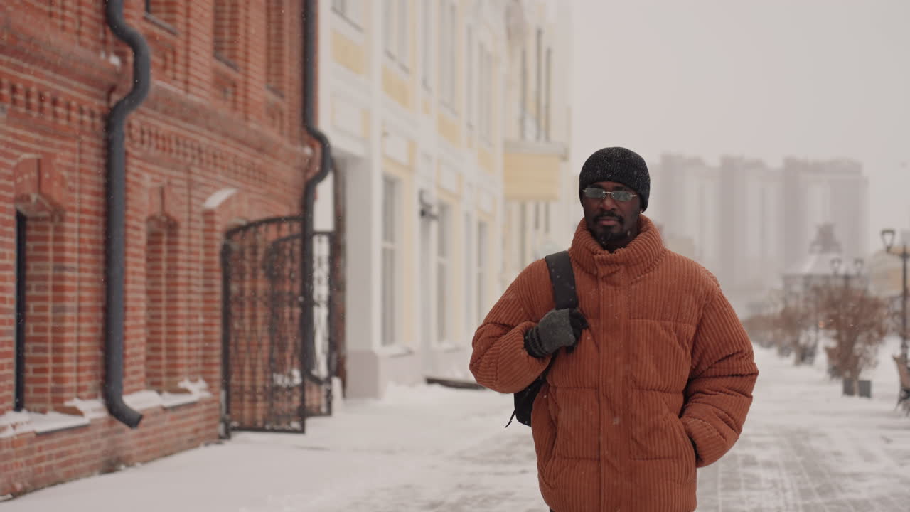 Man walking toward camera snowy boulevard, downtown skyline backdrop, orange puffer jacket, beanie and shoulder bag, direct gaze and subtle smile, closeup frames with visible breath, warm color