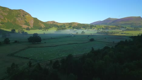 Rise-over-dark-woodland-towards-misted-fields-with-winding-stream-and-mountains-in-the-English-Lake-District,-Cumbria,-UK