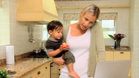 young mother in kitchen coping with laptop phone and baby