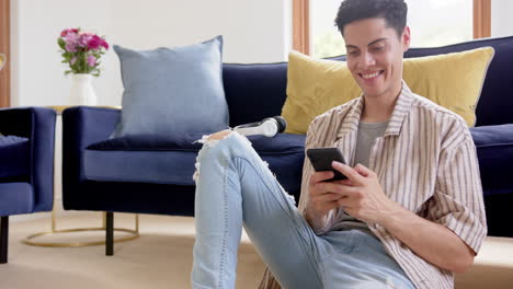 hombre biracial feliz sentado en el suelo usando teléfono inteligente en la sala de estar, espacio de copia, cámara lenta