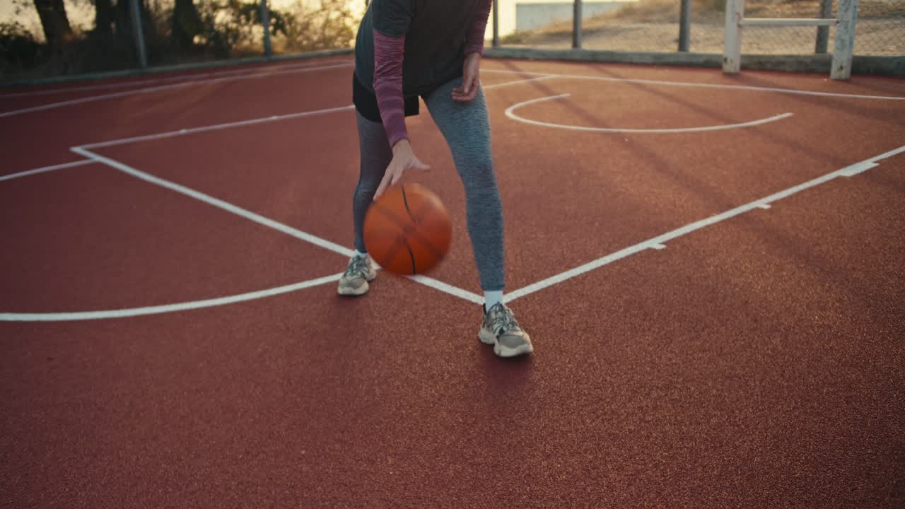 primer plano una chica con un uniforme deportivo golpea una pelota de baloncesto naranja desde un piso rojo en una cancha de la calle al amanecer en verano. la chica se dedica a los deportes y desarrolla la capacidad de jugar al baloncesto por la mañana
