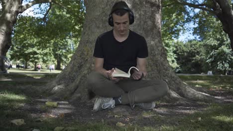 joven blanco sentado bajo un árbol leyendo un libro en un parque en la ciudad de cambridge, inglaterra