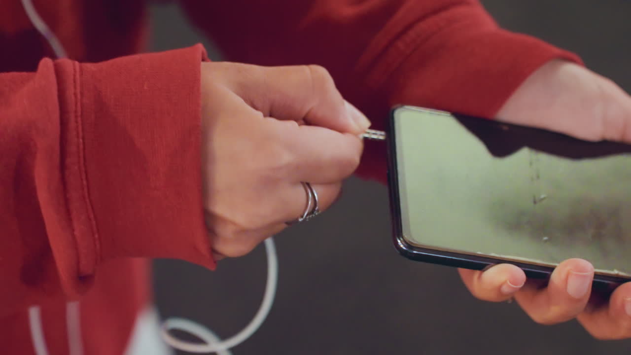 Tight shot hands connecting earbud cable to smartphone, red hoodie sleeves, silver rings, cracked screen reflection, runner prepping playlist for run, looping white cable, city dusk atmosphere