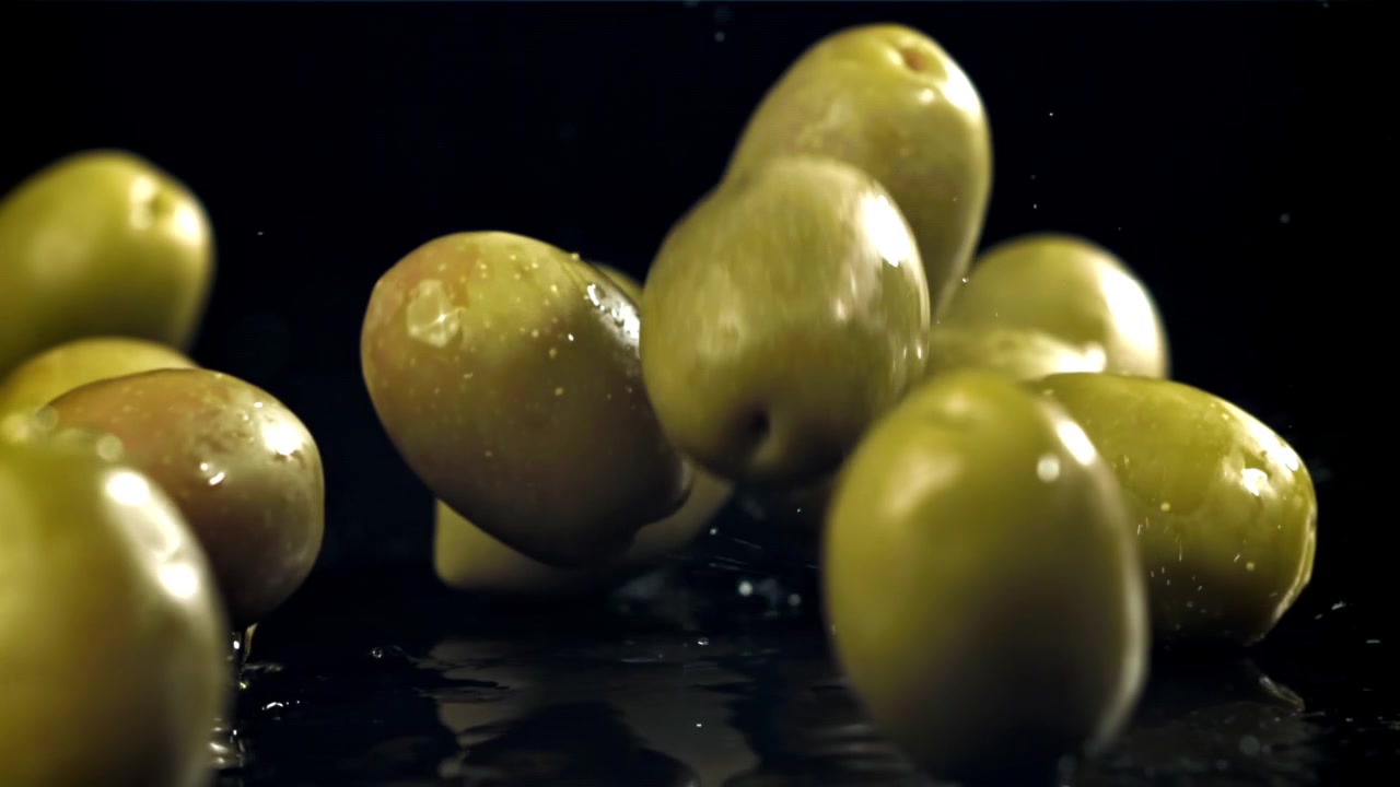 Olive verdi che cadono e schizzano su una superficie nera bagnata al rallentatore.