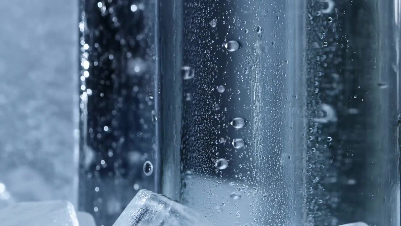 Tight low-angle view of transparent vertical glass cylinder amid irregular ice cubes, showing active bubbles, surface condensation, glossy highlights, and fresh chilled liquid mood