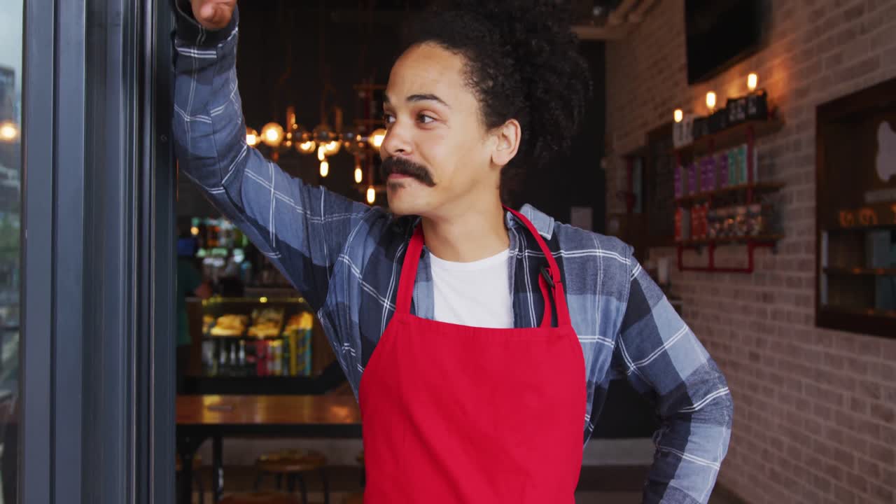 barista de raza mixta con bigote inclinado en la puerta de la cafetería sonriendo