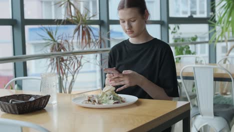 young woman eating a fresh salad at a wooden table in a modern restaurant with large windows, natural light, and urban views. relaxed dining scene